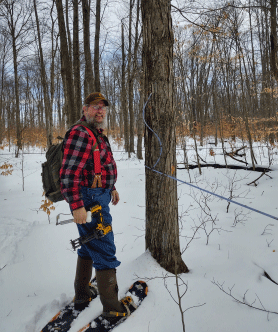 Dave Yeany in the woods, wearing snowshoes and red plaid, carrying sugaring equipment in his backpack.