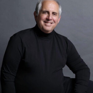Studio picture of a smiling man with grey hair and a black turtleneck.
